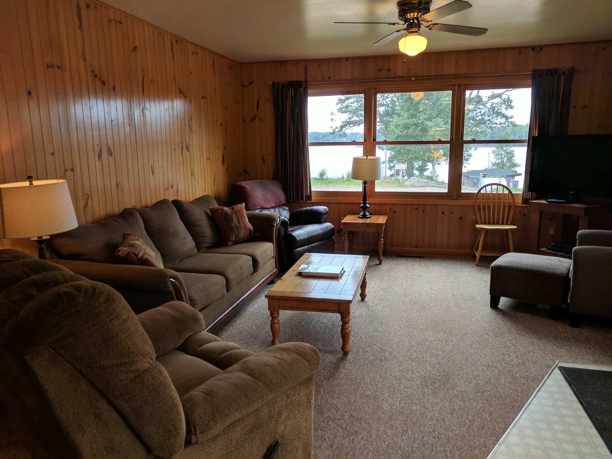 A cozy living room with wood paneling, a brown sofa, armchair, coffee table, and ceiling fan.