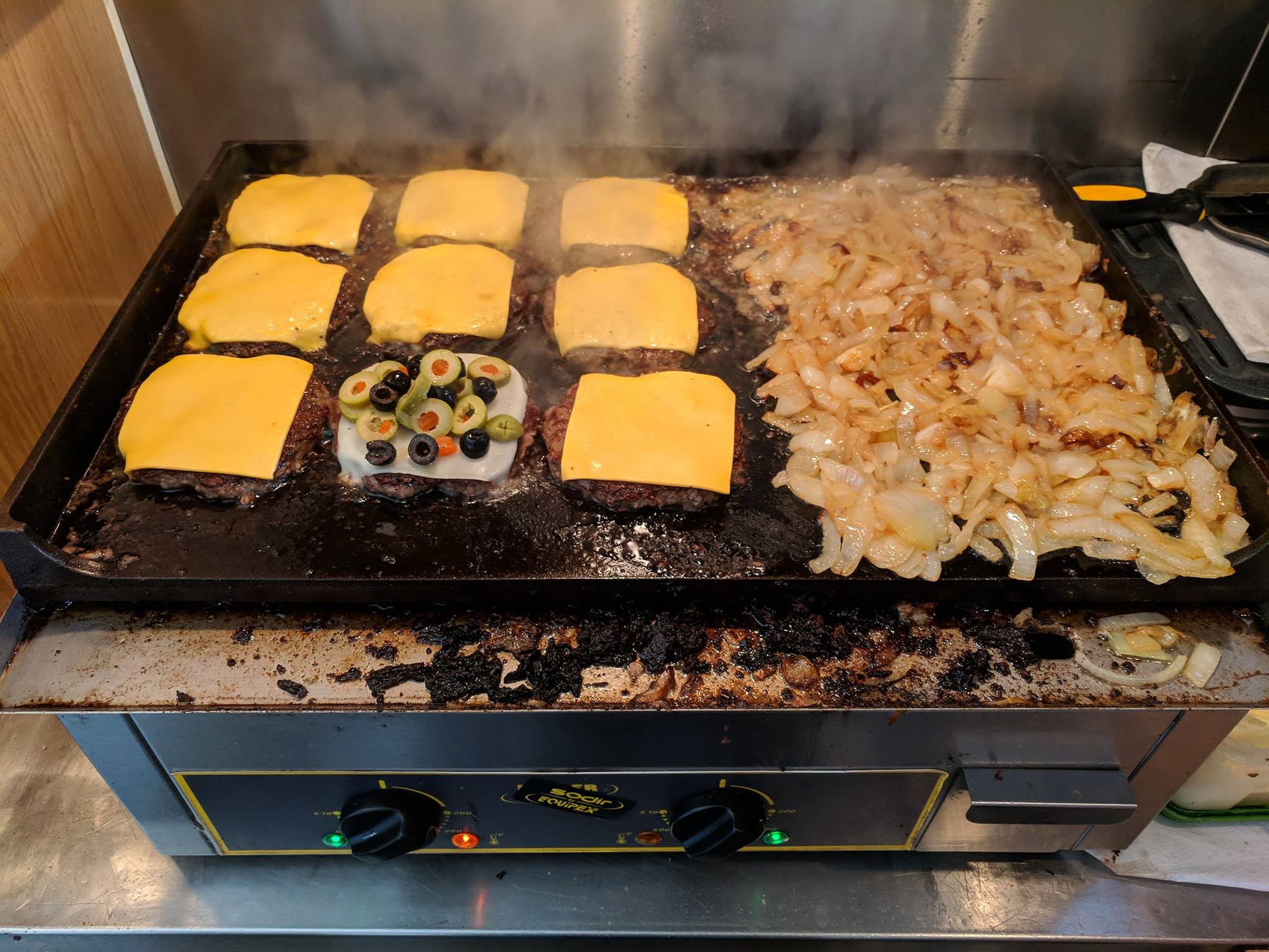 A grill with burgers, cheese, onions, and olives being cooked.