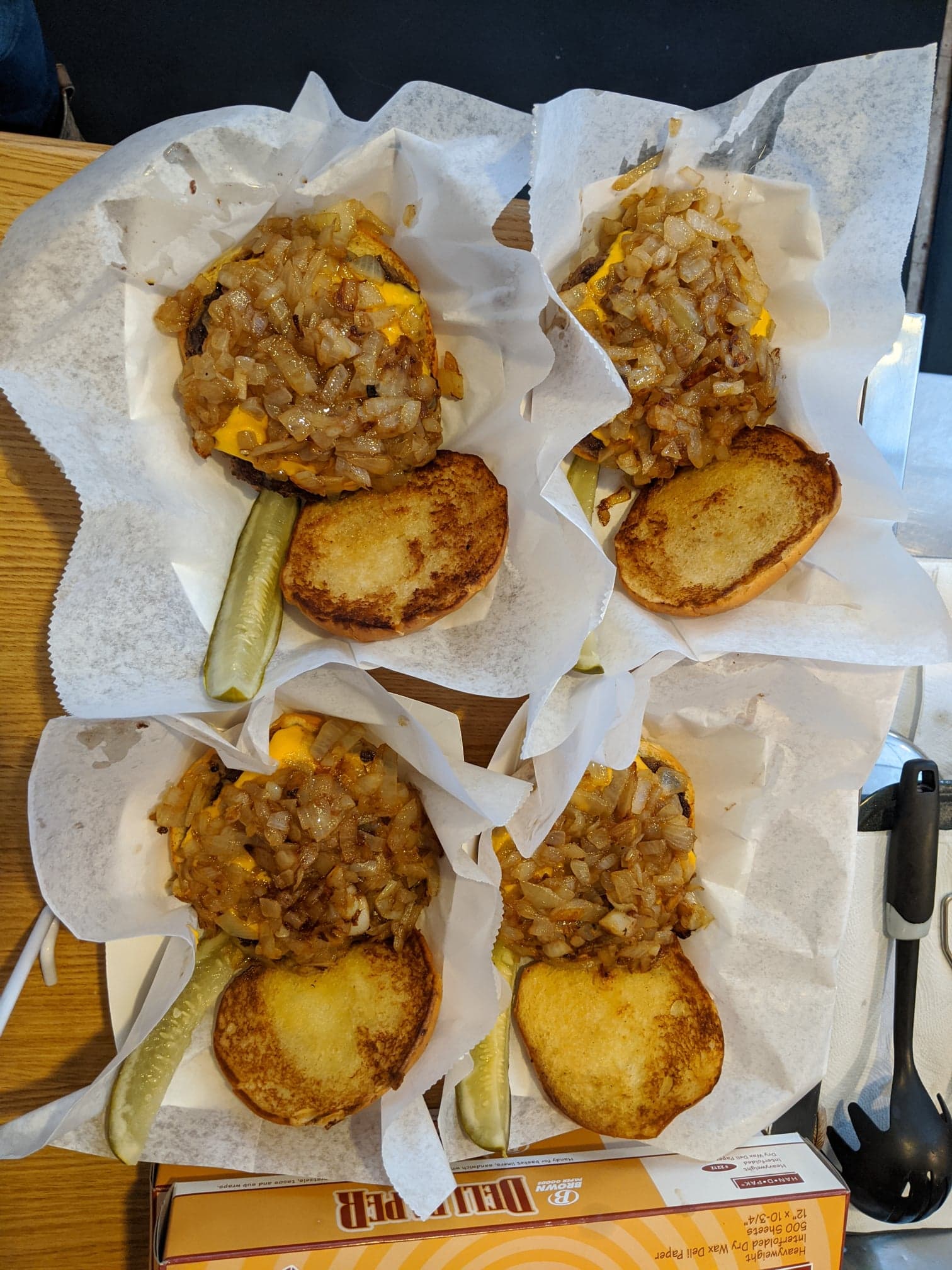 Several burgers with toasted buns and toppings on paper liners, with pickles on the side.