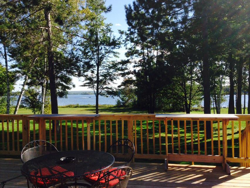 A deck with a wooden railing and metal table and chairs overlooking a lake through trees.